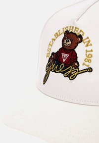 Cappello bianco con orsetto marrone ricamato che indossa una maglietta rossa, testo dorato "Fondato nel 1981" e scritta "guess" in oro.