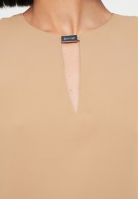 Μπεζ φόρεμα με ντεκολτέ τύπου σχισμής και ασημένια ετικέτα Calvin Klein ως λεπτομέρεια. Το ύφασμα είναι λείο και ελαφρύ.