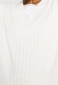 Maglione a coste color crema con scollatura a V. Presenta un tessuto testurizzato con leggere variazioni, indossato sopra una camicia bianca.