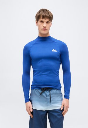 Mladý muž v priliehavom modrom rash garde s dlhým rukávom a modrých plavkách s prechodom farby a čiernou šnúrkou na stiahnutie, stojaci pred čistým pozadím.