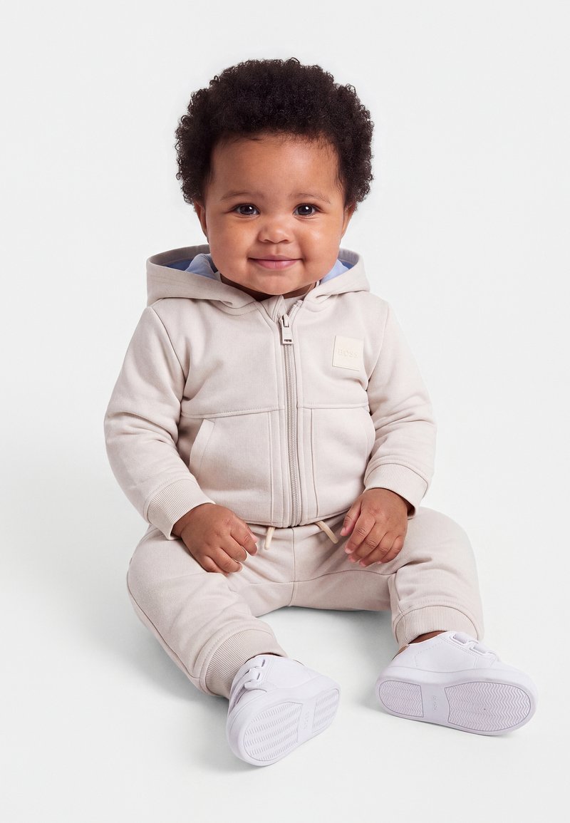 Bébé souriant aux cheveux bouclés portant une veste à capuche beige clair, un pantalon assorti et des chaussures blanches, assis sur un fond uni.
