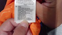 Oranžika värvi jakikate silt, mis näitab materjale: 96% taaskasutatud polüester, 4% spandex, 100% polüuretaan, hooldusjuhised ja taaskasutusteave.