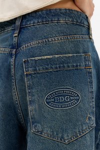 Nærbilde av en baklomme på blå denimjeans med sydd oval BDG-logo-patch og frynsete detaljer på toppen.