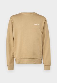 Beige sweatshirt met geribde ronde hals en manchetten, met subtiele "Club1990" tekst in het wit op de linker borst. Soepele textuur.