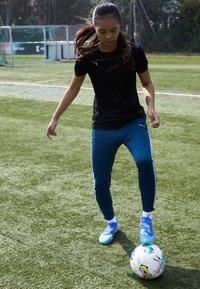 Camiseta atlética negra, pantalones ajustados azules con rayas blancas y zapatos de fútbol azules. Un balón de fútbol está colocado cerca de los pies del jugador.