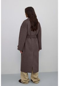 Brązowy wełniany płaszcz z paskiem w talii, szerokimi rękawami i rozcięciem z tyłu. Połączony z beżowymi spodniami o szerokich nogawkach i brązowym obuwiem.