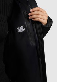 Doudoune noire avec un extérieur ribbed et un intérieur brillant. Comprend une poche zippée avec un patch de la marque à l'intérieur.