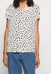 Top beige a maniche corte con motivo maculato nero, scollatura rotonda e vestibilità rilassata, abbinato a pantaloni in denim.