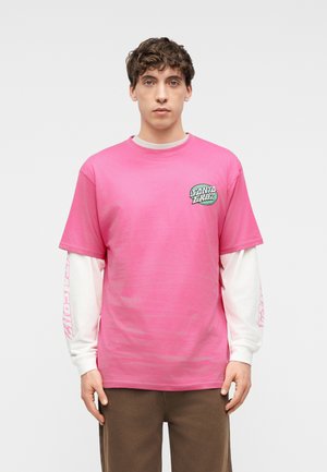 Νεαρός άνδρας με κοντά σγουρά μαλλιά, φορώντας ροζ κοντομάνικη μπλούζα Santa Cruz πάνω από λευκό μακρυμάνικο πουκάμισο και καφέ παντελόνι, στέκεται ακίνητος.