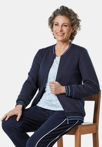 Marineblauwe gewatteerde bomberjack met contrasterende blauwe accenten, gecombineerd met een lichtblauw bloemen T-shirt en marineblauwe broek met witte zijkanten.