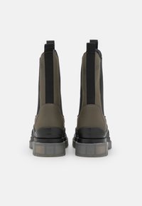 Botas de tobillo en verde oliva con paneles laterales elásticos negros, una suela de goma translúcida y gruesa, y detalles cosidos en el talón.