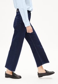 Jeans de pierna ancha de color azul oscuro, de mezclilla, con corte recto, dos bolsillos traseros y un dobladillo limpio. Combinados con mocasines negros.