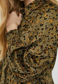 Grön blus med en samlad ärm och ett flerfärgat prickmönster i brunt, guld och svart. Texturerad tyg med krage.