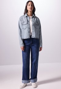 Giacca di jeans azzurro chiaro con due tasche sul petto, indossata su una camicia bianca. Jeans a gamba larga blu scuro, risvoltati alla caviglia, e sneakers bianche.