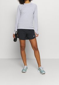 Vaaleanharmaa pitkähihainen urheilupaita, jossa on pyöreä kaula-aukko, yhdessä mustien shortsien kanssa, joissa on valkoinen Nike-logo, ja vaaleansiniset lenkkikengät.