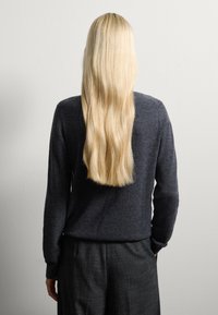 Pull noir long avec une texture douce, associé à un pantalon noir à motifs. De longs cheveux blonds raides sont visibles.