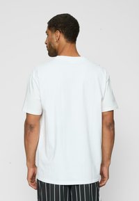 Tricou alb din bumbac, cu mâneci scurte, decolteu rotund, textură netedă și croială lejeră, purtat cu pantaloni raiati. Vedere din spate.