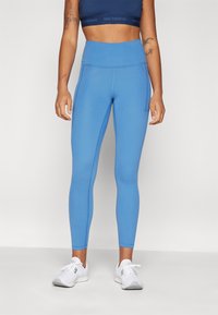 Niebieskie legginsy z wysokim stanem, wykonane z gładkiego, elastycznego materiału z bocznymi kieszeniami. Noszone z białymi butami sportowymi.