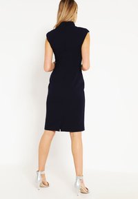Granatowa dopasowana sukienka z rękawami w kształcie kaptura, wymodelowanymi szwami, rozcięciem z tyłu i wysokim dekoltem. W zestawieniu ze srebrnymi sandałami na wysokim obcasie.
