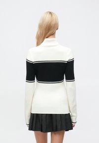 Blond kvinne sett bakfra, iført en hvit høyhalset genser med svart horisontal stripe og et plissert mørkt skjørt.