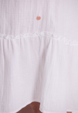 Weißes Baumwollkleid aus strukturiertem Stoff, mit einem Rüschensaum und einem kleinen, markierten Metallanhänger nahe der oberen Mitte.