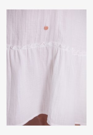 Weißes Baumwollkleid aus strukturiertem Stoff, mit einem Rüschensaum und einem kleinen, markierten Metallanhänger nahe der oberen Mitte.