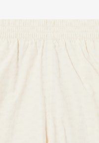 Light cream shorts with an elastic waistband, featuring a subtle all-over logo pattern. Soft material, smooth texture, and loose fit.
