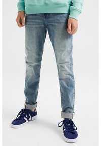 Jeans de mezclilla azul claro con un ajuste ajustado, textura deslavada y puños enrollados, combinados con zapatillas de ante azules sobre un fondo blanco.
