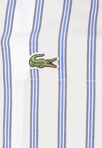Tessuto bianco con righe verticali blu e grigie, caratterizzato da un logo di coccodrillo ricamato in verde con un accentuazione rossa.