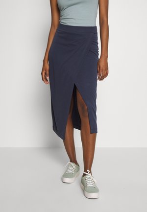 Jupe portefeuille bleu marine avec une fente haute, fabriquée en tissu doux. Associée à un haut vert clair et des baskets grises. Texture lisse et design simple.