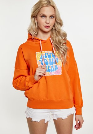 Ung kvinde med langt blondt hår iført orange hættetrøje med farverigt "Live Your Dream"-tryk og hvide distressed shorts.