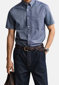 Homme portant une chemise bleue à manches courtes boutonnée et un jean foncé avec une ceinture tressée marron, tenant une veste en cuir marron dans une main.