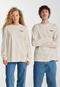 Helle beige Langarmshirts mit einem kleinen Logo auf der Brust, kombiniert mit lockeren blauen Jeans. Der Stoff scheint weich und angenehm zu sein.