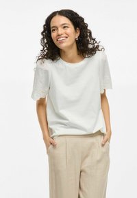 Camiseta blanca de manga corta con detalles de encaje en las mangas, combinada con pantalones beige. La tela parece suave y tiene un corte relajado.