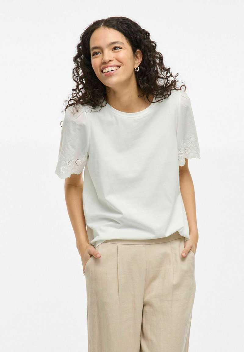 Camiseta blanca de manga corta con detalles de encaje en las mangas, combinada con pantalones beige. La tela parece suave y tiene un corte relajado.
