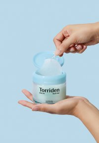 Klar krukke med et lyseblåt låg, der indeholder en multifunktionel hudpleje pad. Etiketten lyder "Torriden DIVE IN Multi Pad." En hånd holder krukken.