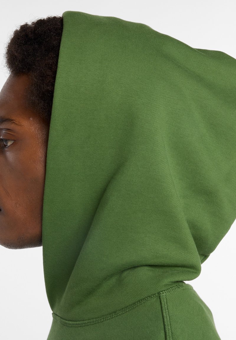 Sweat à capuche vert avec une texture douce et des détails côtelés, ajusté autour du visage, avec des coutures visibles le long de l'ourlet.