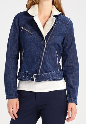 Kvinde iført en blå denimjakke med hvid shearling-krave, asymmetrisk lynlås, lynlåslommer og bælte i taljen.