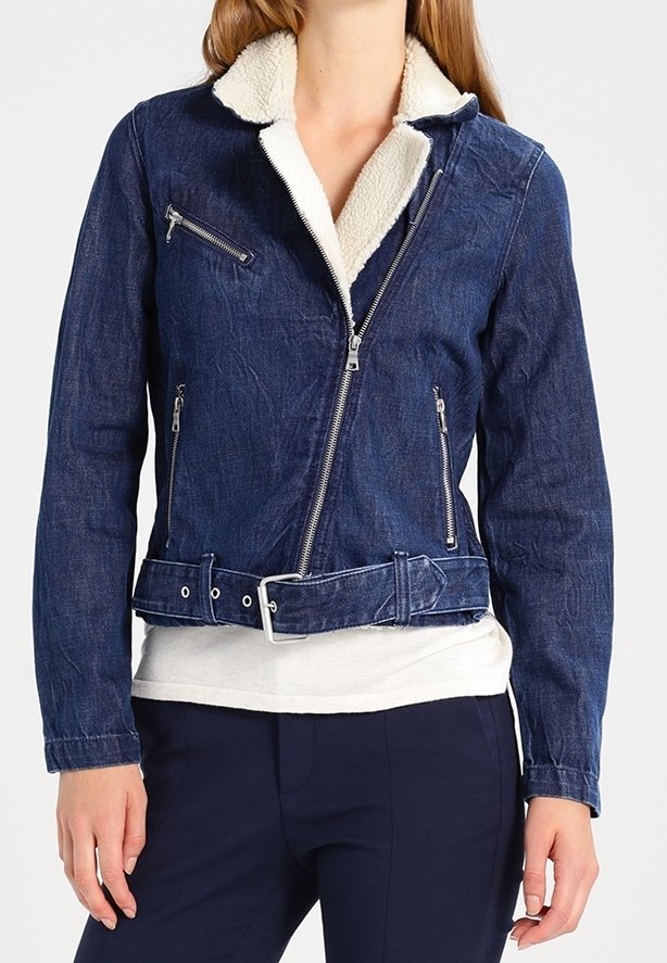 Femme portant une veste en jean bleue avec col en shearling blanc, fermeture éclair asymétrique, poches zippées et ceinture à la taille.