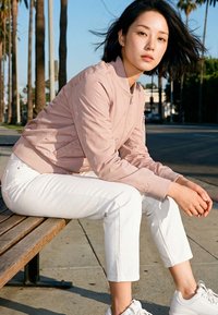 Woman with black hair wearing a light pink jacket, white pants, and white sneakers sitting on a wooden bench outdoors near palm trees.