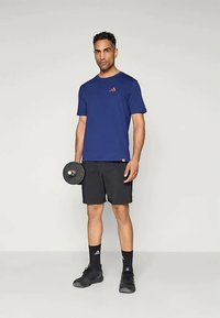 Homem vestido com uma camiseta atlética azul-marinho e calções pretos, segurando um haltere, em pé contra um fundo claro e liso.