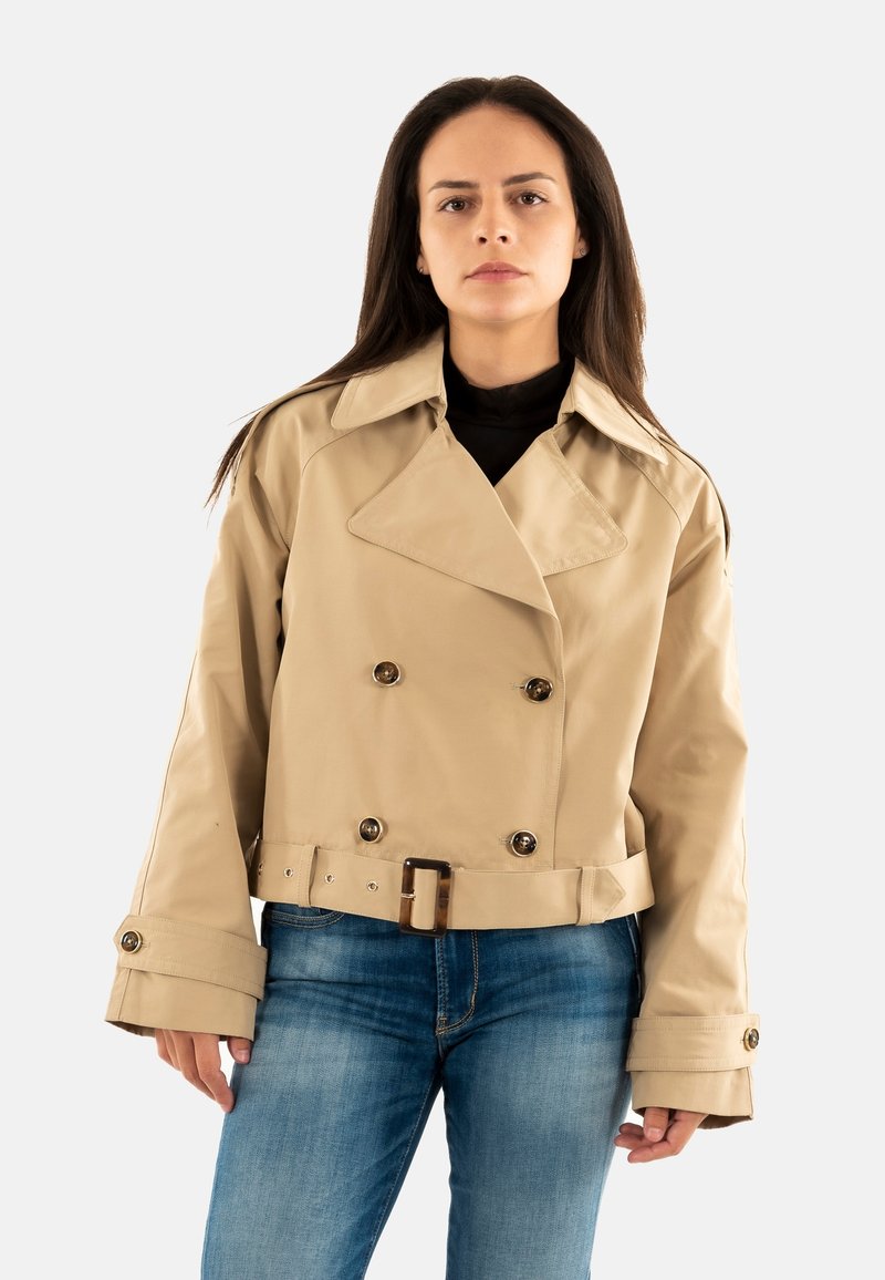 Donna con lunghi capelli scuri che indossa un trench corto doppiopetto beige con cintura e jeans blu, in piedi contro uno sfondo bianco.