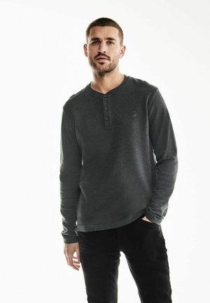 Camisa henley de mangas largas en gris oscuro con un escote redondeado, abertura de tres botones en el pecho y un sutil detalle del logo en el lado izquierdo.