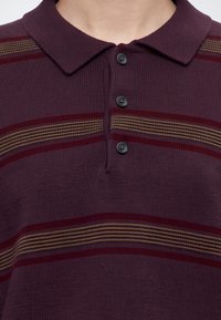 Gros plan sur la poitrine d'une personne portant un polo en maille bordeaux avec une patte de trois boutons et des rayures horizontales beige et rouge foncé.
