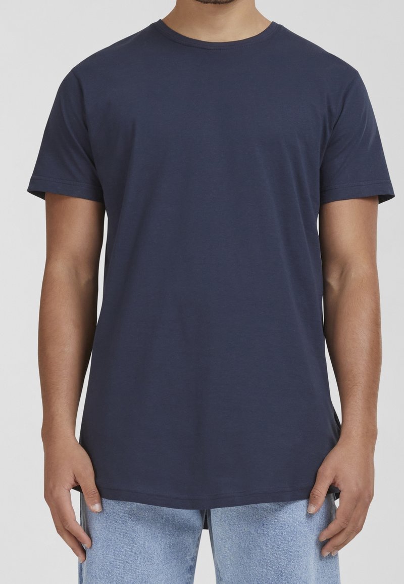Homme portant un t-shirt uni bleu marine à manches courtes et un jean bleu clair, se tenant avec les bras détendus le long du corps.