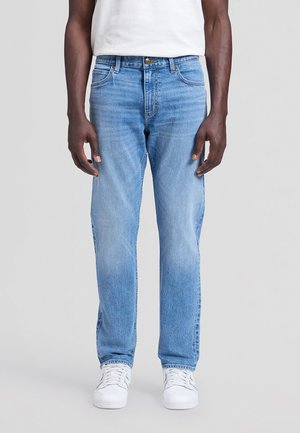 Personne portant un jean droit bleu clair, des baskets blanches et une chemise blanche, debout devant un fond gris clair uni.