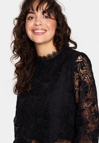 Blusa nera con pizzo in alto, caratterizzata da motivi floreali, collo alto con bordi scallop e maniche lunghe trasparenti. Tessuto morbido con dettagli intricati.