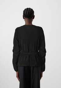Haut noir plissé à manches longues avec une ceinture nouée à la taille. Présente un col rond et une surface texturée. Design simple et élégant.