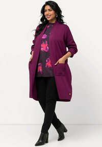Langer, offener Cardigan aus reichhaltigem lila Stoff mit gerippter Textur, ausgestattet mit zwei vorderen Taschen und kombiniert mit einem floral gemusterten Oberteil und schwarzen Hosen.