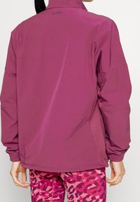 Chaqueta morada con un alto cuello, tejido texturizado, paneles laterales de malla y puños elásticos, que presenta un logo en la parte posterior del cuello.